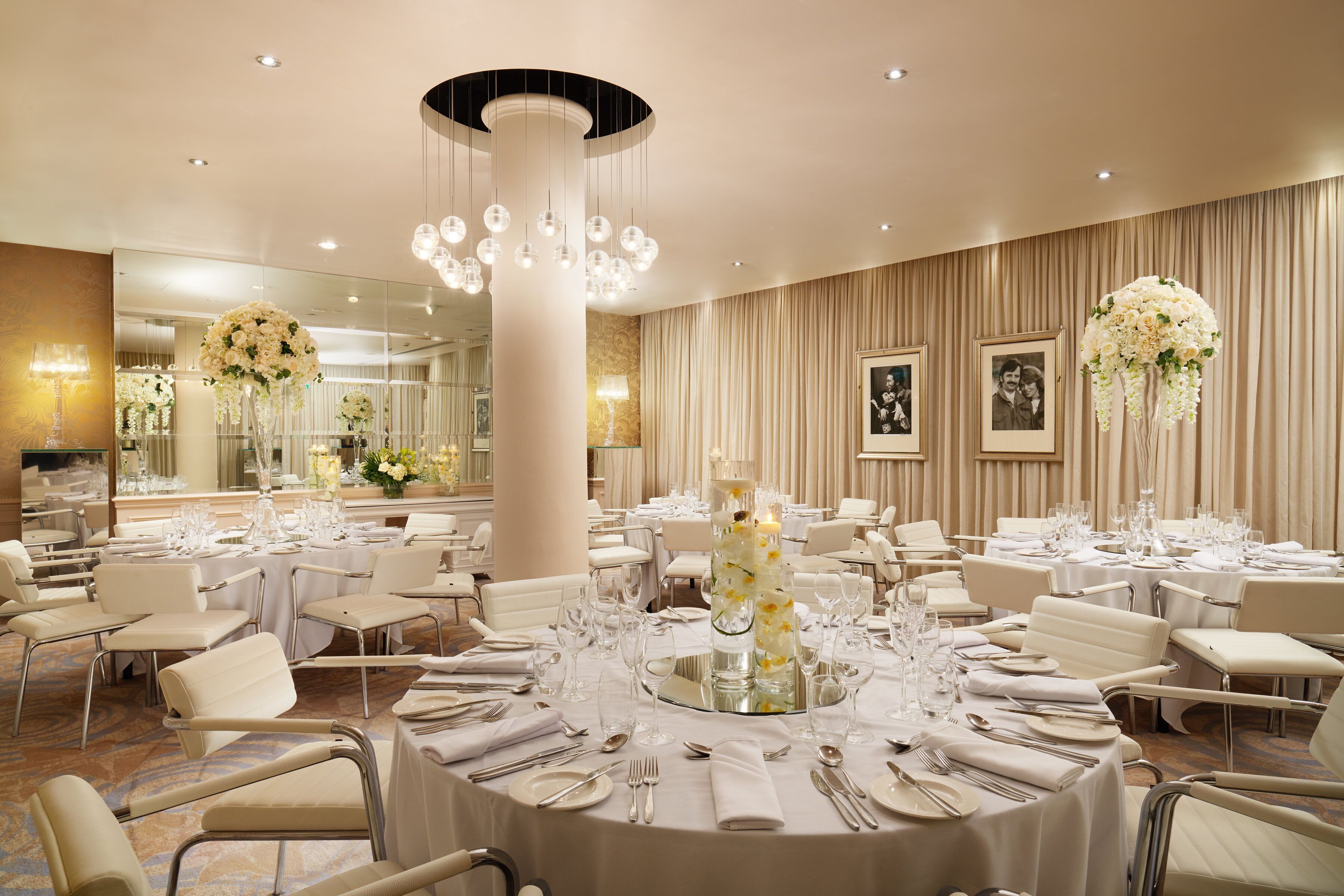 Un salón de banquetes con mesas redondas dispuestas para comer, una columna decorativa central y arreglos florales colgantes.