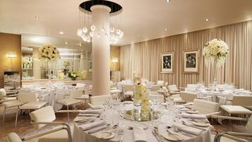 A banquet hall with round tables set for dining, a central decorative column, and hanging floral arrangements.
