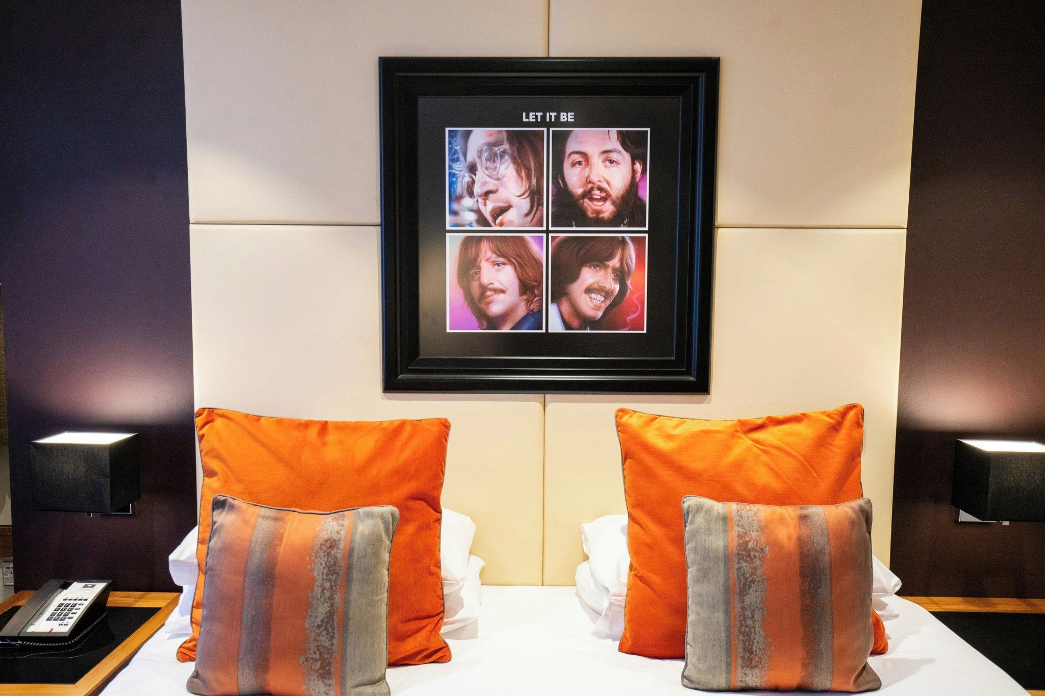 A hotel room with a framed picture on the wall, two orange pillows, a bedside table with a phone, and a wall-mounted lamp.