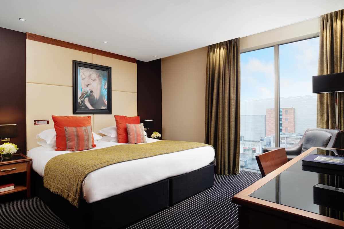 A hotel room with a large bed, a desk with a chair, a view of the city through large windows, and a framed picture on the wall.