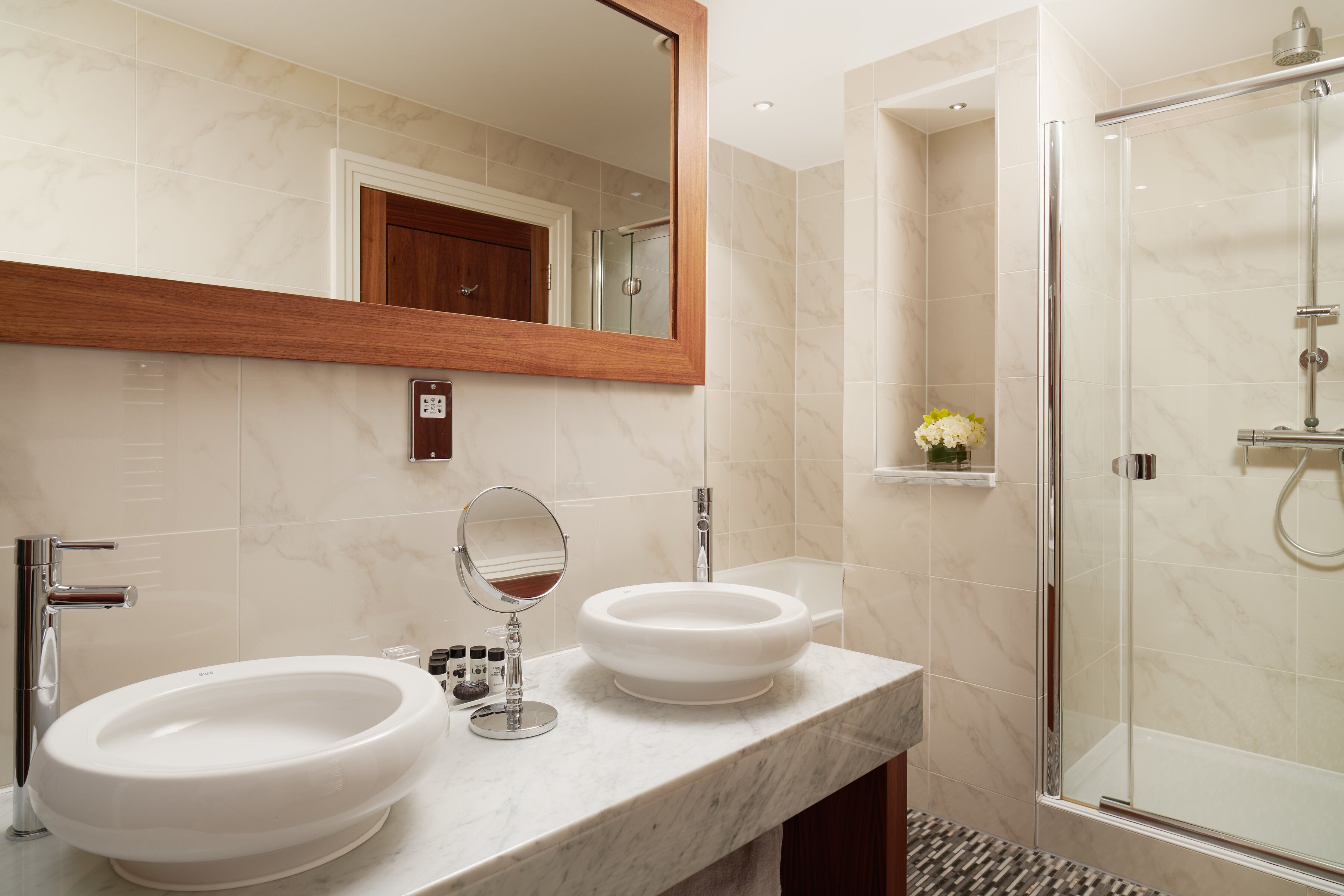 A bathroom with two sinks, a mirror, a shower, and a marble-tiled wall.