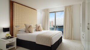 A modern hotel room with a large bed, a bedside table with a vase of flowers, and a view of the city through large windows. - Hard Days Night Hotel Liverpool (Liverpool)
