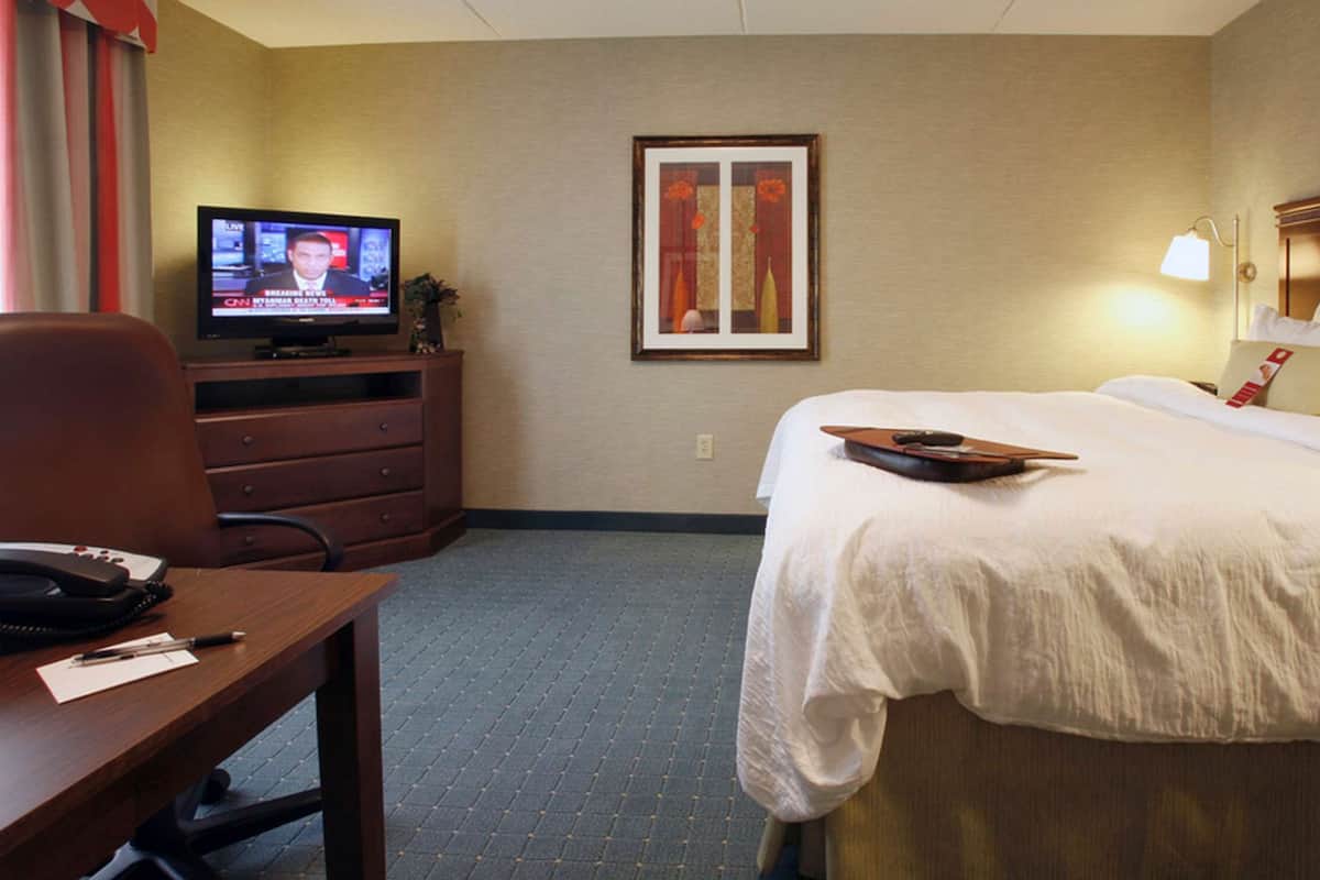 Une chambre d’hôtel avec un lit, un bureau avec un téléphone et des documents, un téléviseur diffusant les actualités et un tableau encadré au mur.