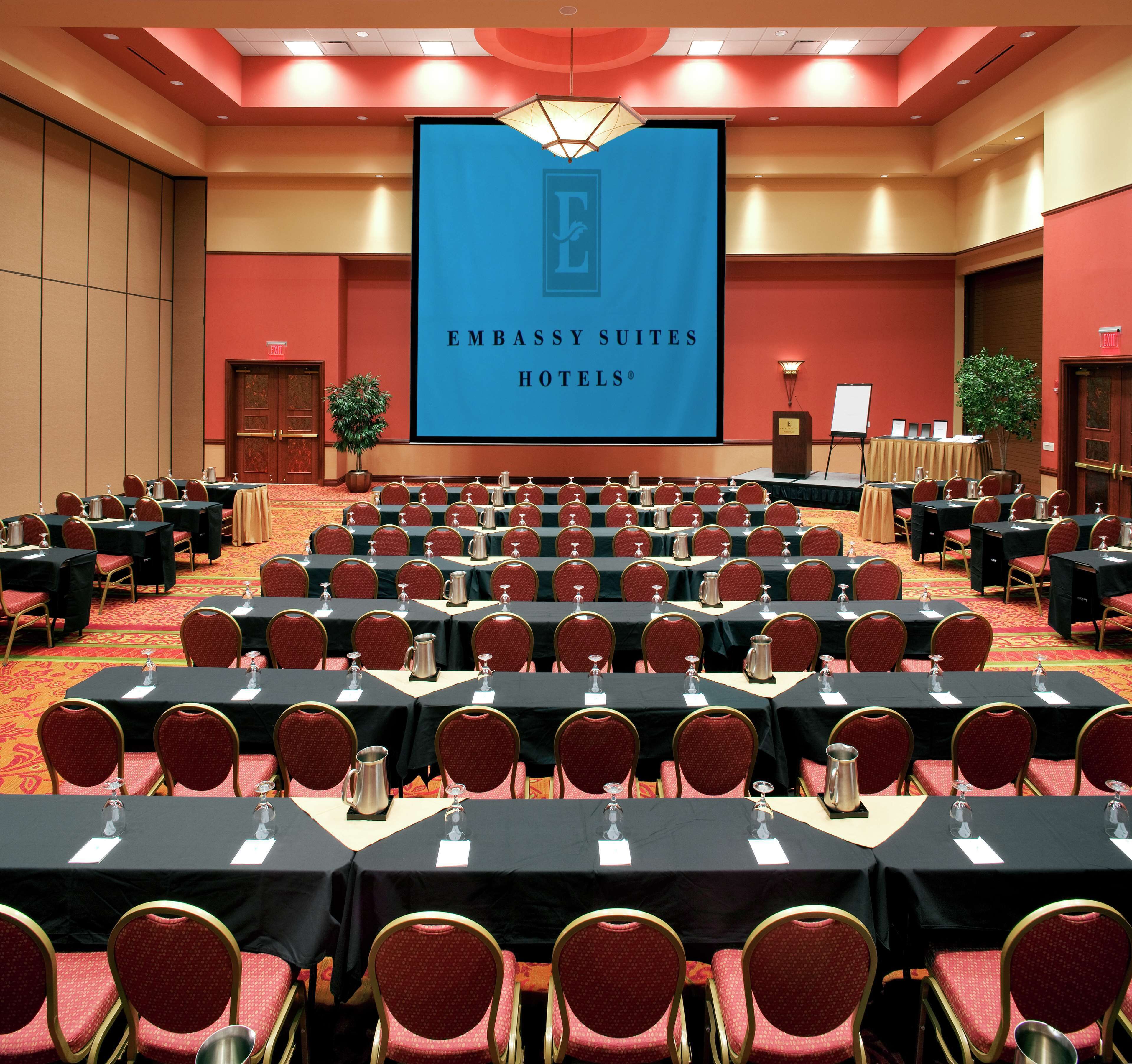 A conference room with rows of tables and chairs, a large screen displaying 'EMBASSY SUITES HOTELS', and a podium with a microphone.