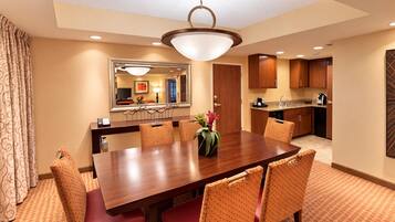 A hotel dining area with a long wooden table, red upholstered chairs, a large mirror, and a kitchen in the background.
