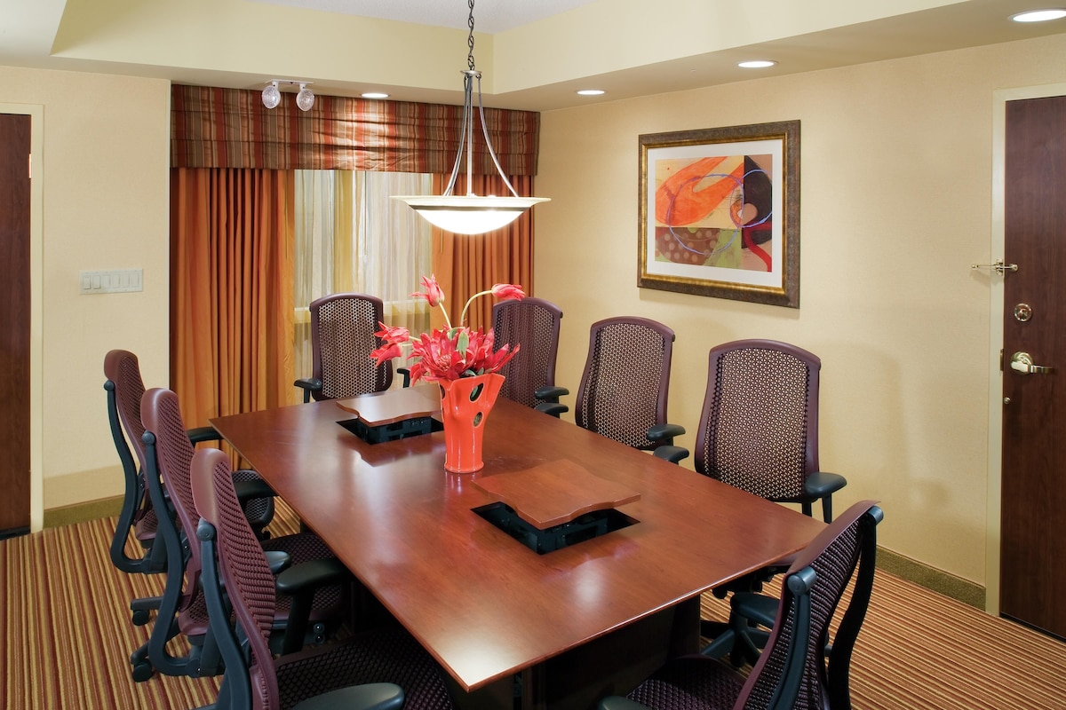 A conference room with a long table, six chairs, a vase with flowers, and a painting on the wall.