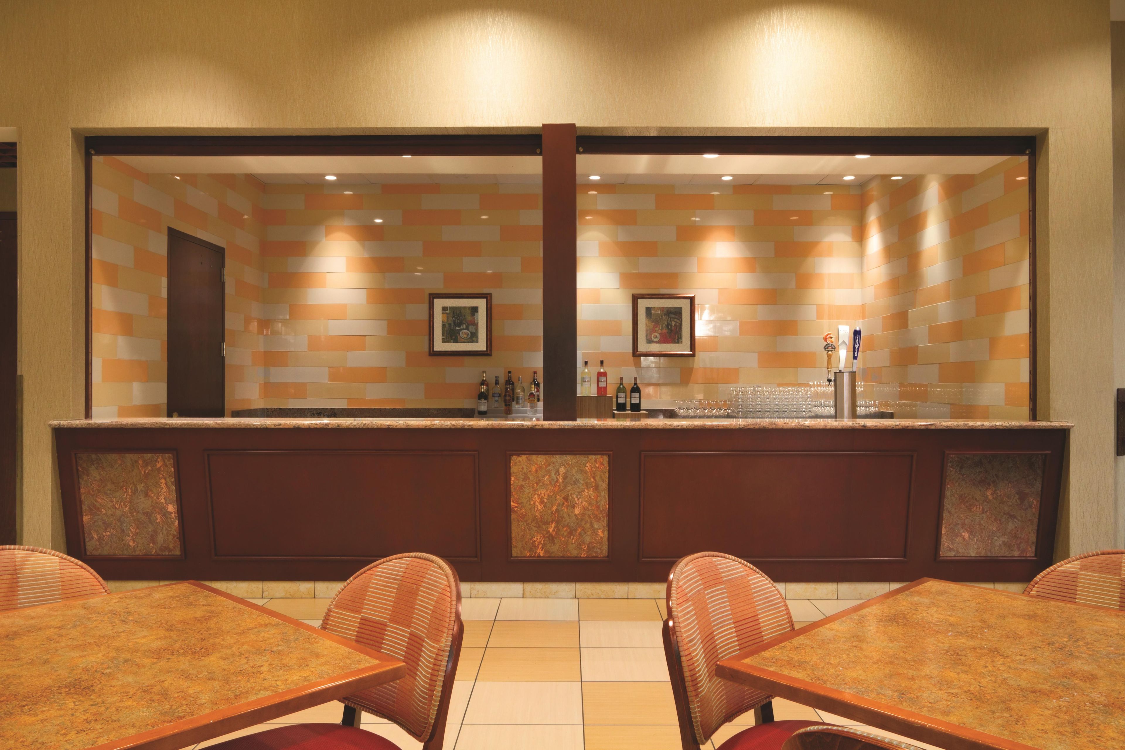 A bar area with a tiled wall, two framed pictures, and a counter with bottles. There are four wicker chairs and a table in front.