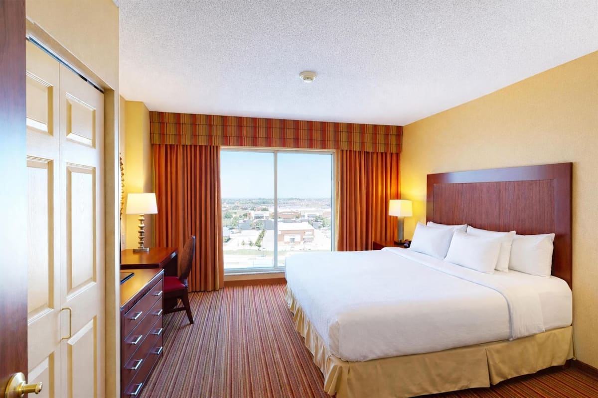 A hotel room with a large bed, a desk with a chair, a lamp, and a view of the city through the window.