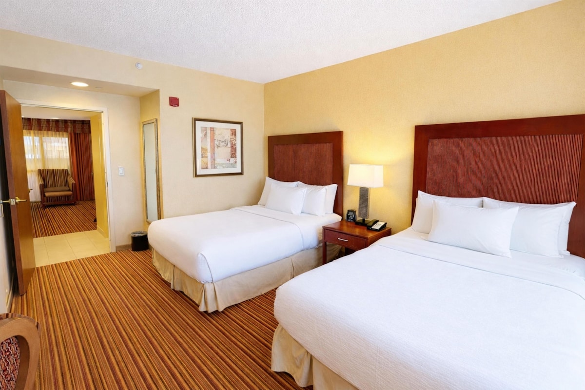A hotel room with two beds, a nightstand with a lamp, a framed picture on the wall, and a carpeted floor.
