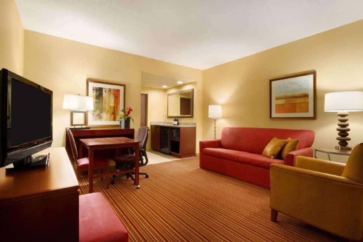 A hotel room with a television, a dining area, a red sofa, a small armchair, and a painting on the wall.