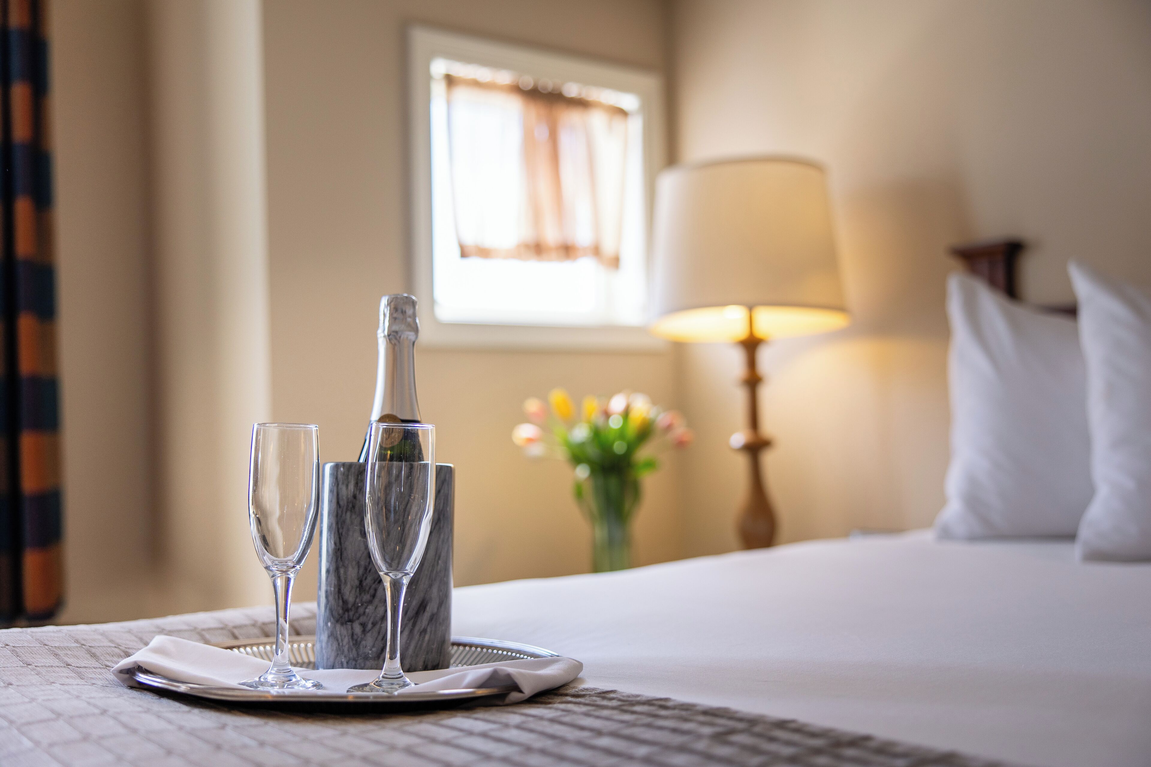 A neatly arranged hotel room with a bed, a bedside lamp, a vase with flowers, and a table with a bottle and glasses.