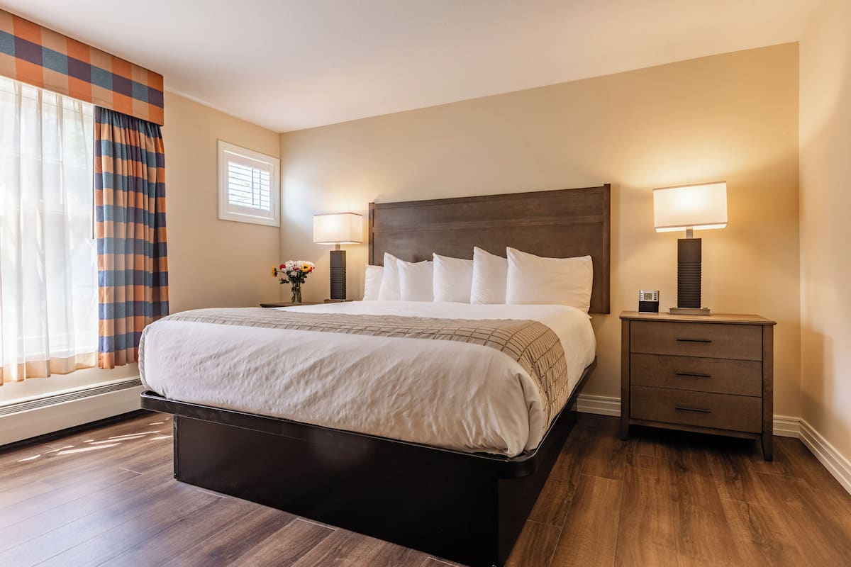 A hotel room with a large bed, two bedside tables with lamps, a window with curtains, and a wall-mounted heater.