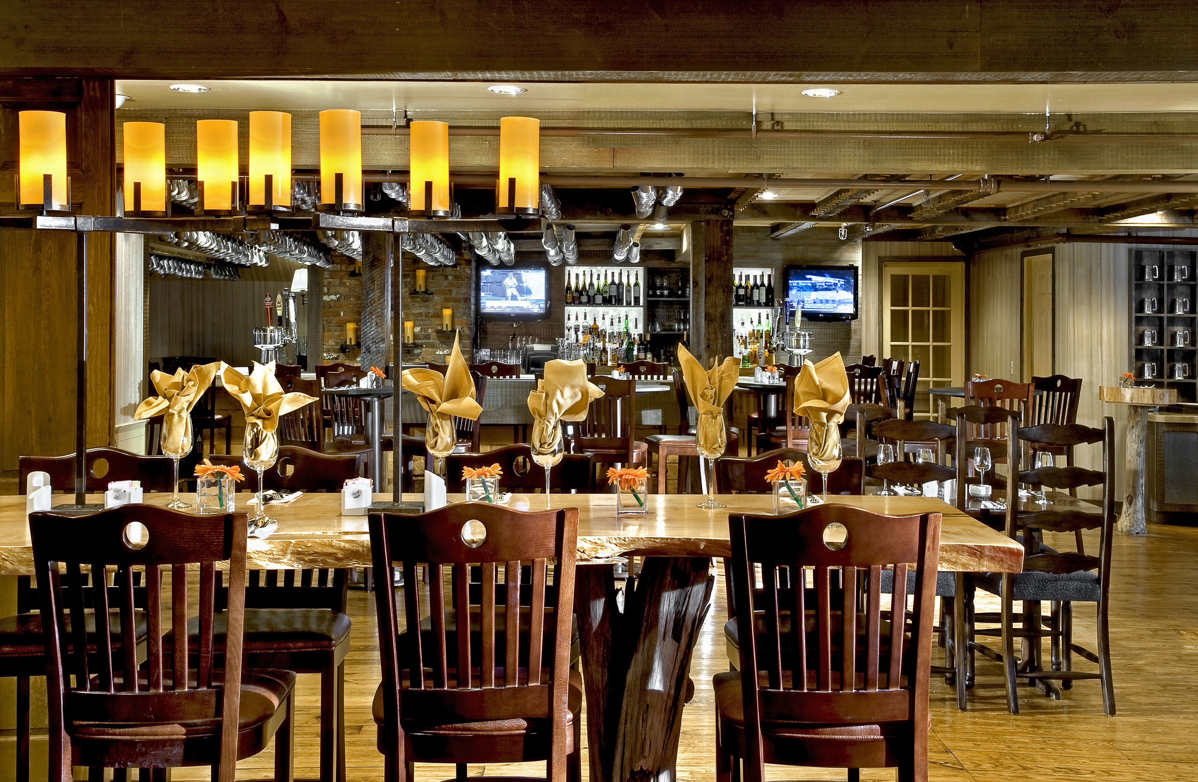 A restaurant with wooden tables and chairs, candles, and a bar area with bottles and a TV screen.