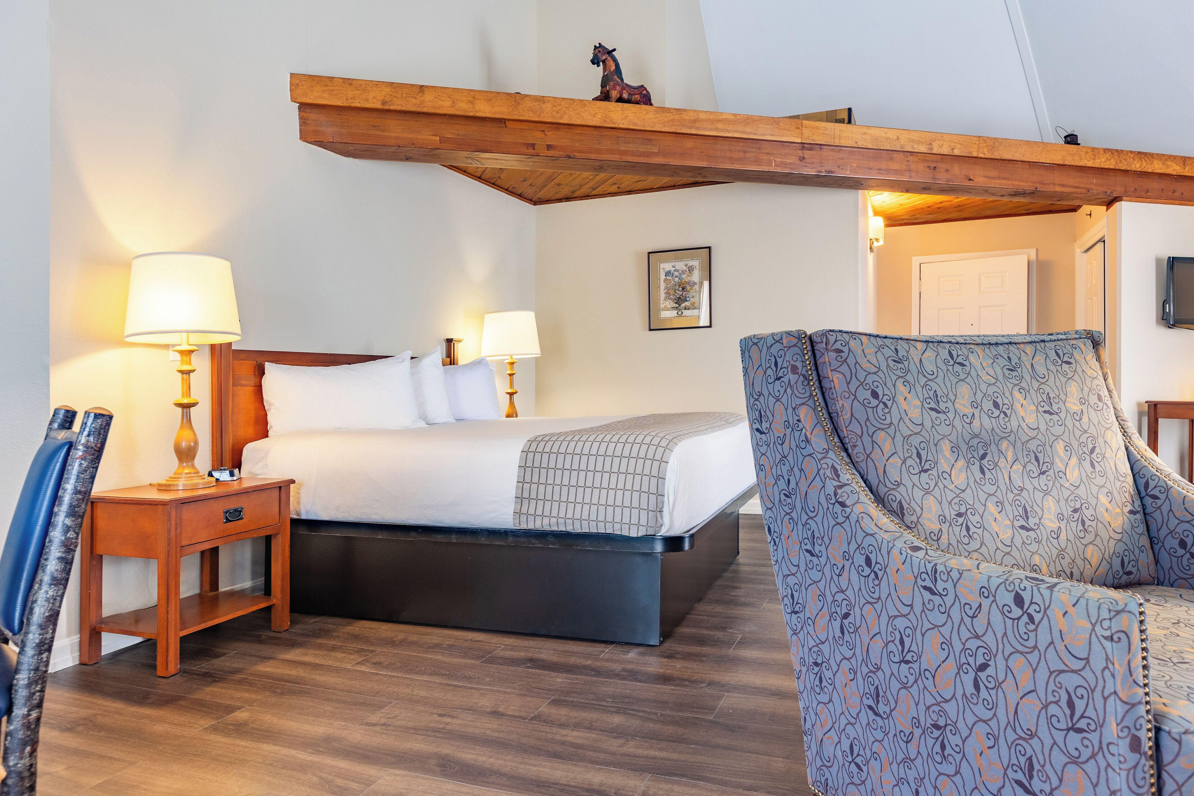 Quarto de hotel com cama, abajures, uma cadeira, cabeceira de madeira, quadro na parede e televisão.