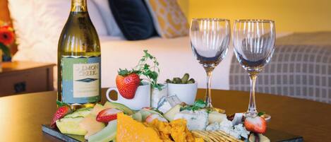 A breakfast platter with various cheeses, fruits, and crackers on a slate board, accompanied by a bottle of wine and two glasses.