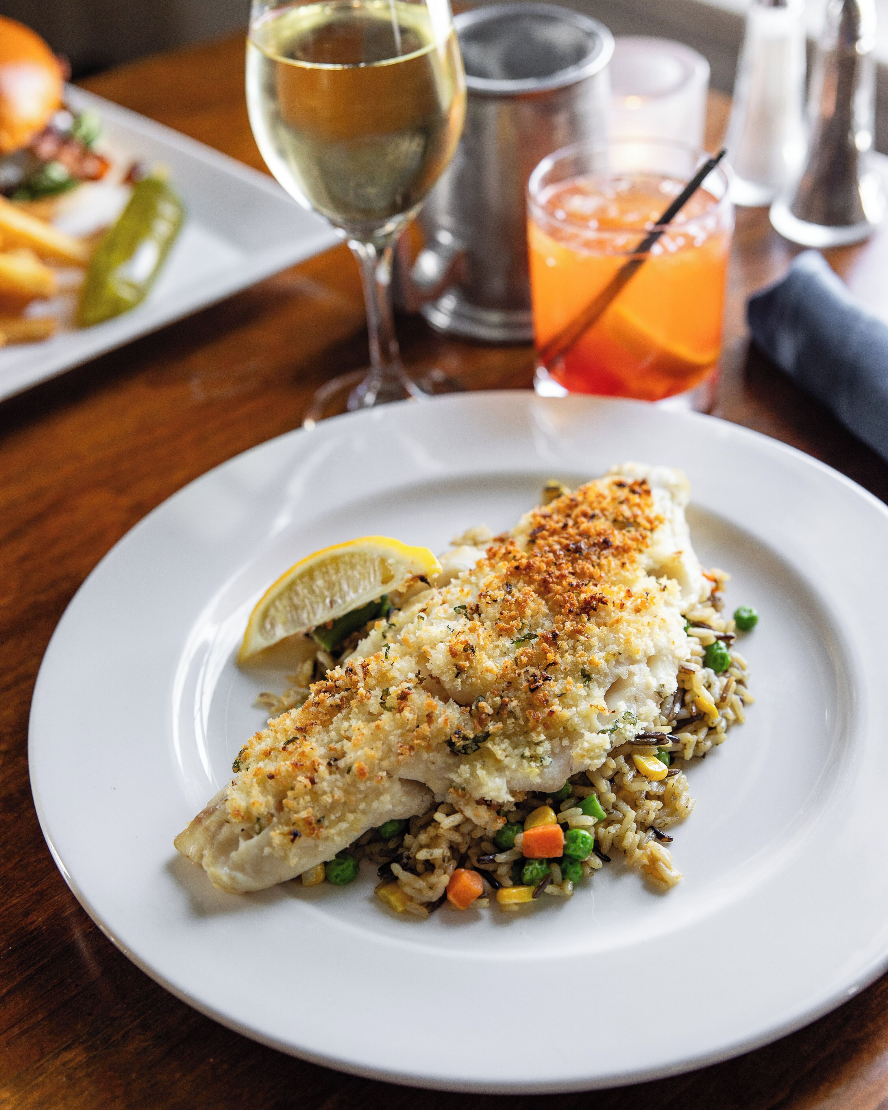 A plate of fish with rice and vegetables, a glass of white wine, and a glass of orange juice on a wooden table.