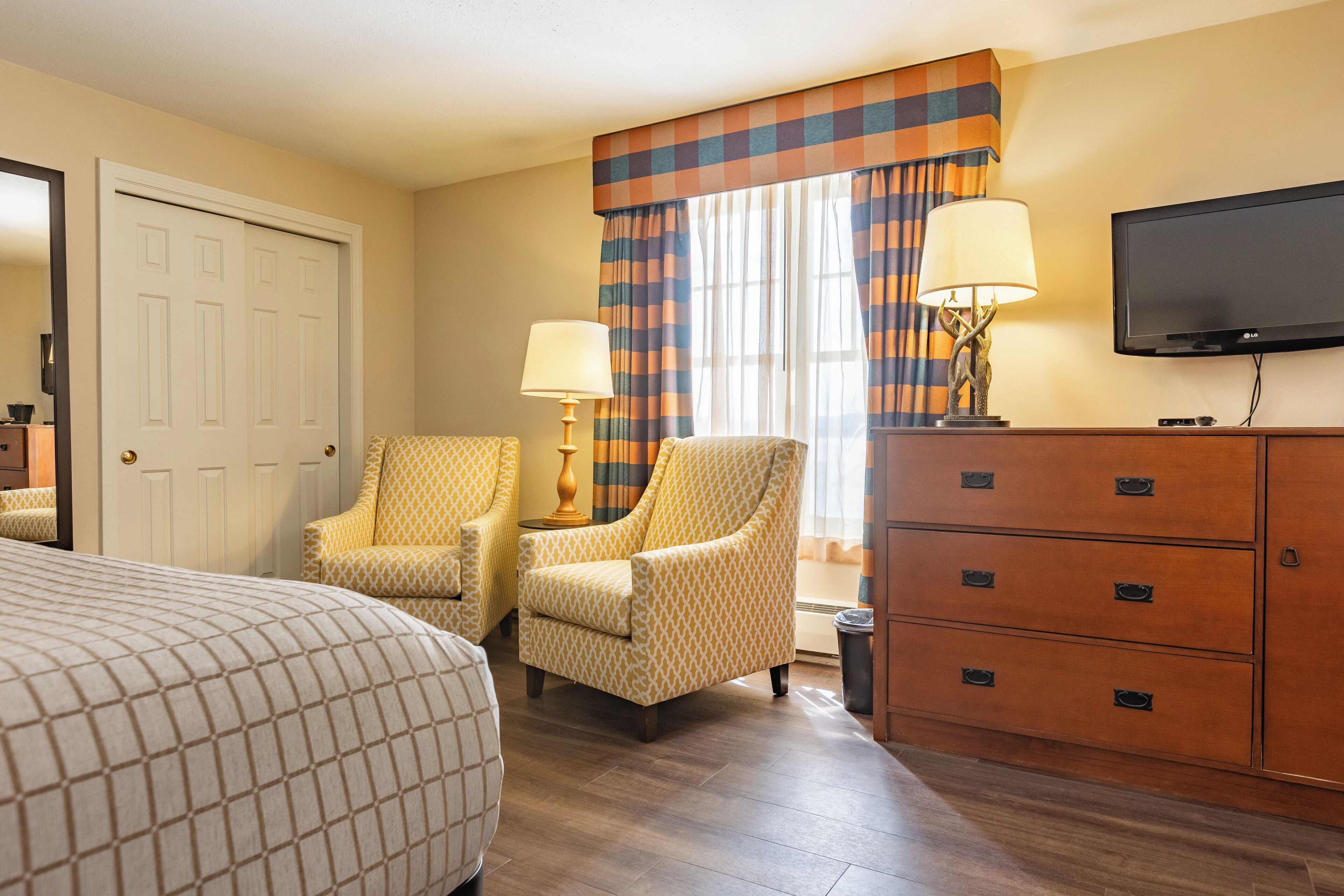 A hotel room with a bed, two armchairs, a dresser, a television, and a window with curtains.
