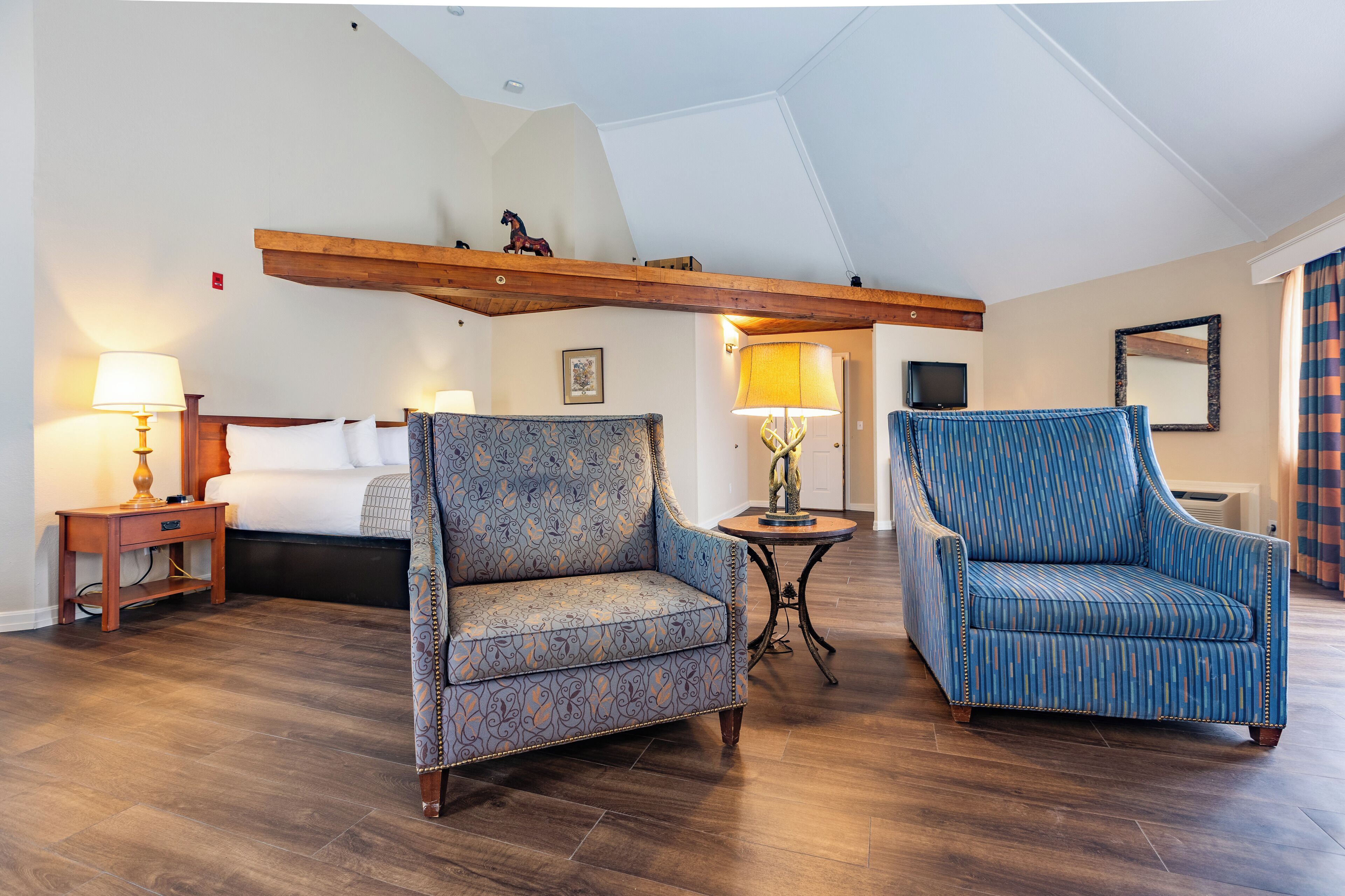 A hotel room with a bed, two patterned armchairs, a nightstand with a lamp, a small table with a lamp, and a wooden beam ceiling.