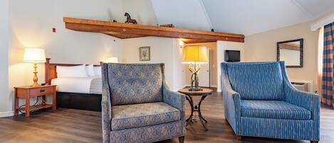 A hotel room with a bed, two patterned armchairs, a nightstand with a lamp, a small table with a lamp, and a wooden beam ceiling.