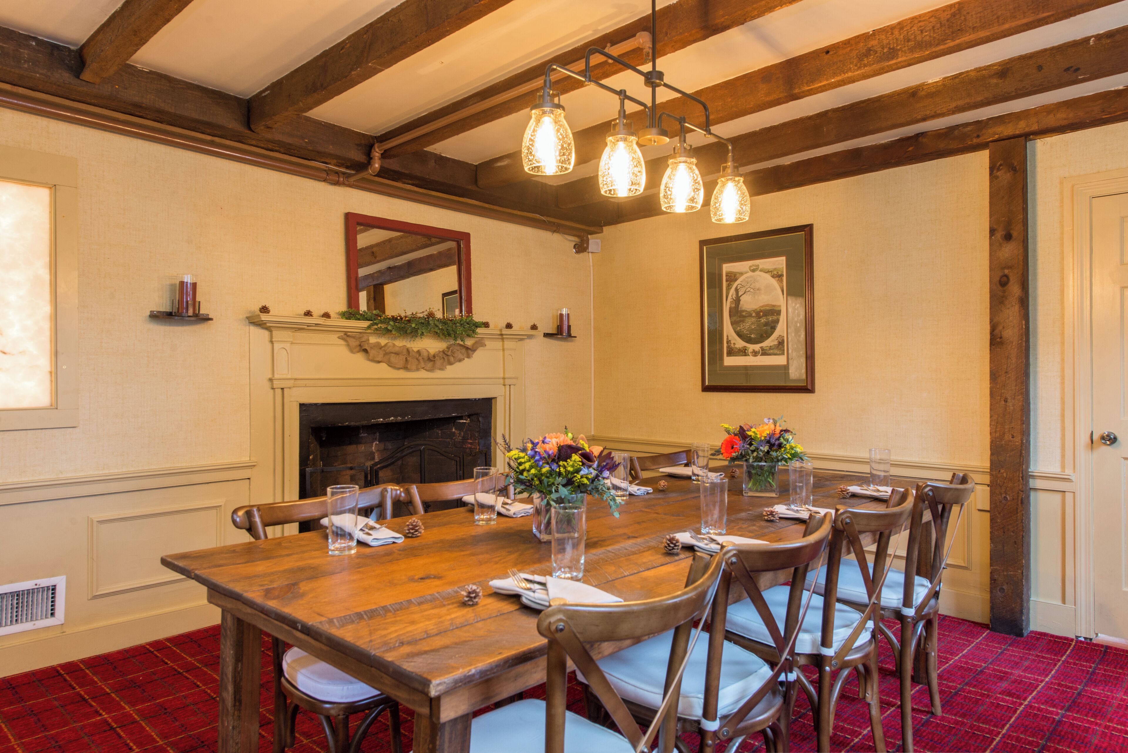 A dining area with a wooden table, chairs, and a fireplace. There are hanging lights above the table and a framed picture on the wall.