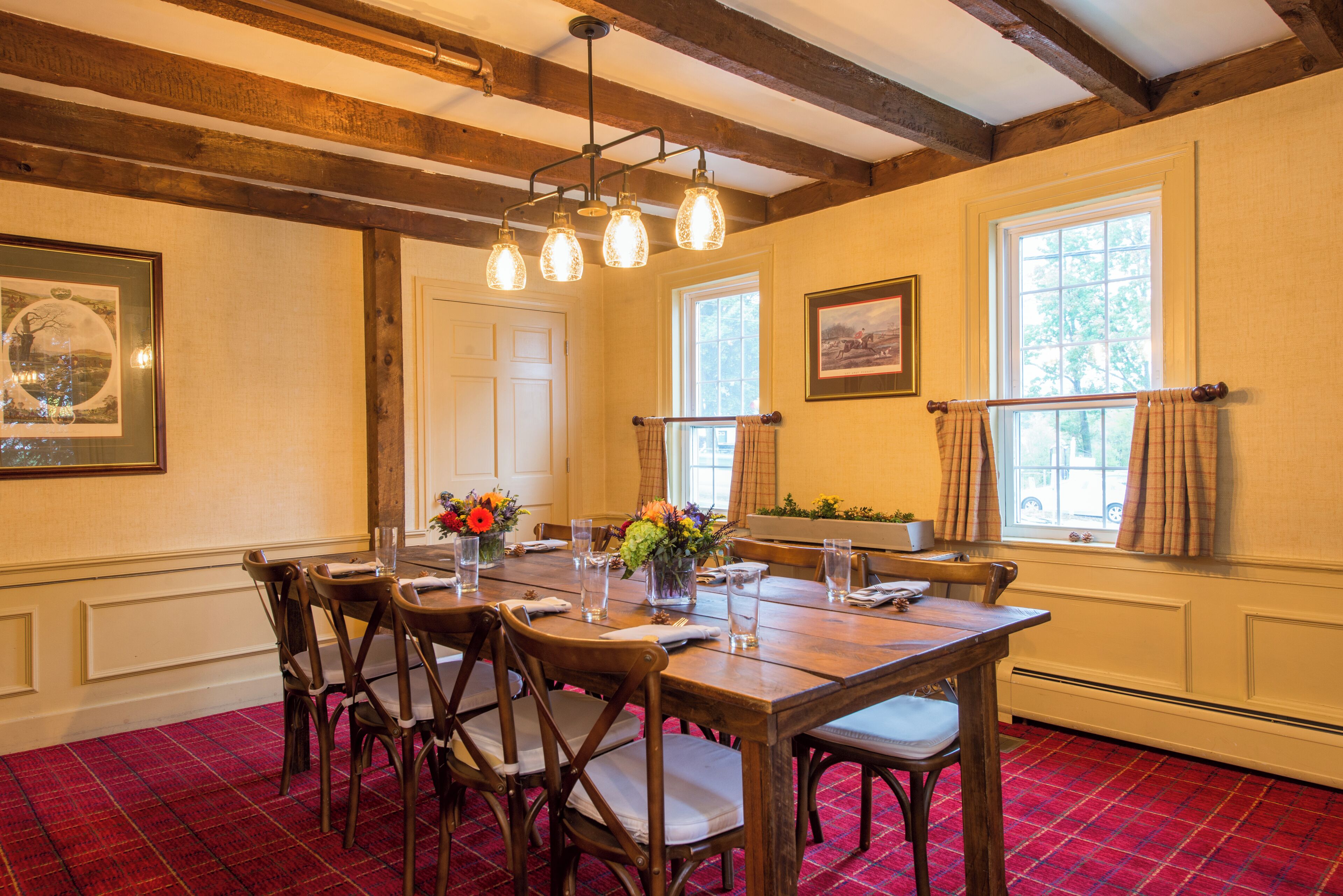 A dining room with a wooden table set for six, surrounded by chairs. There are framed pictures on the walls, a window with curtains, and a red carpeted floor.