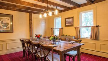 A dining room with a wooden table set for six, surrounded by chairs. There are framed pictures on the walls, a window with curtains, and a red carpeted floor.