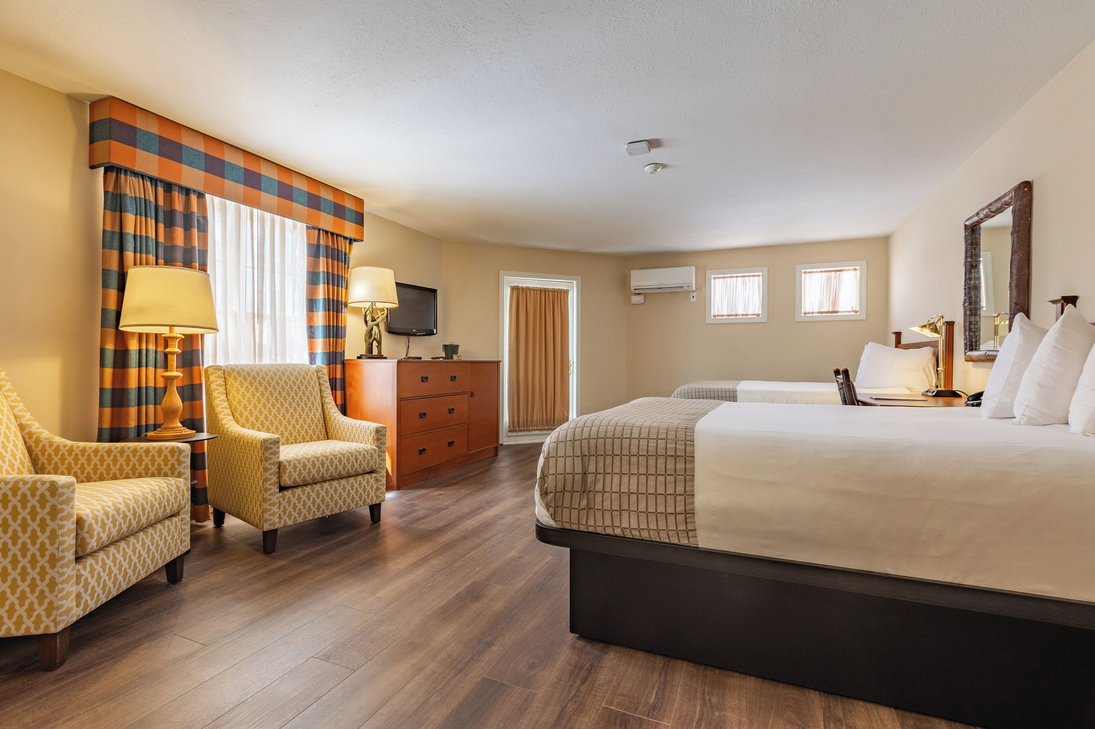 Une chambre d’hôtel avec un grand lit, deux fauteuils, une commode en bois, un téléviseur et une fenêtre avec des rideaux.