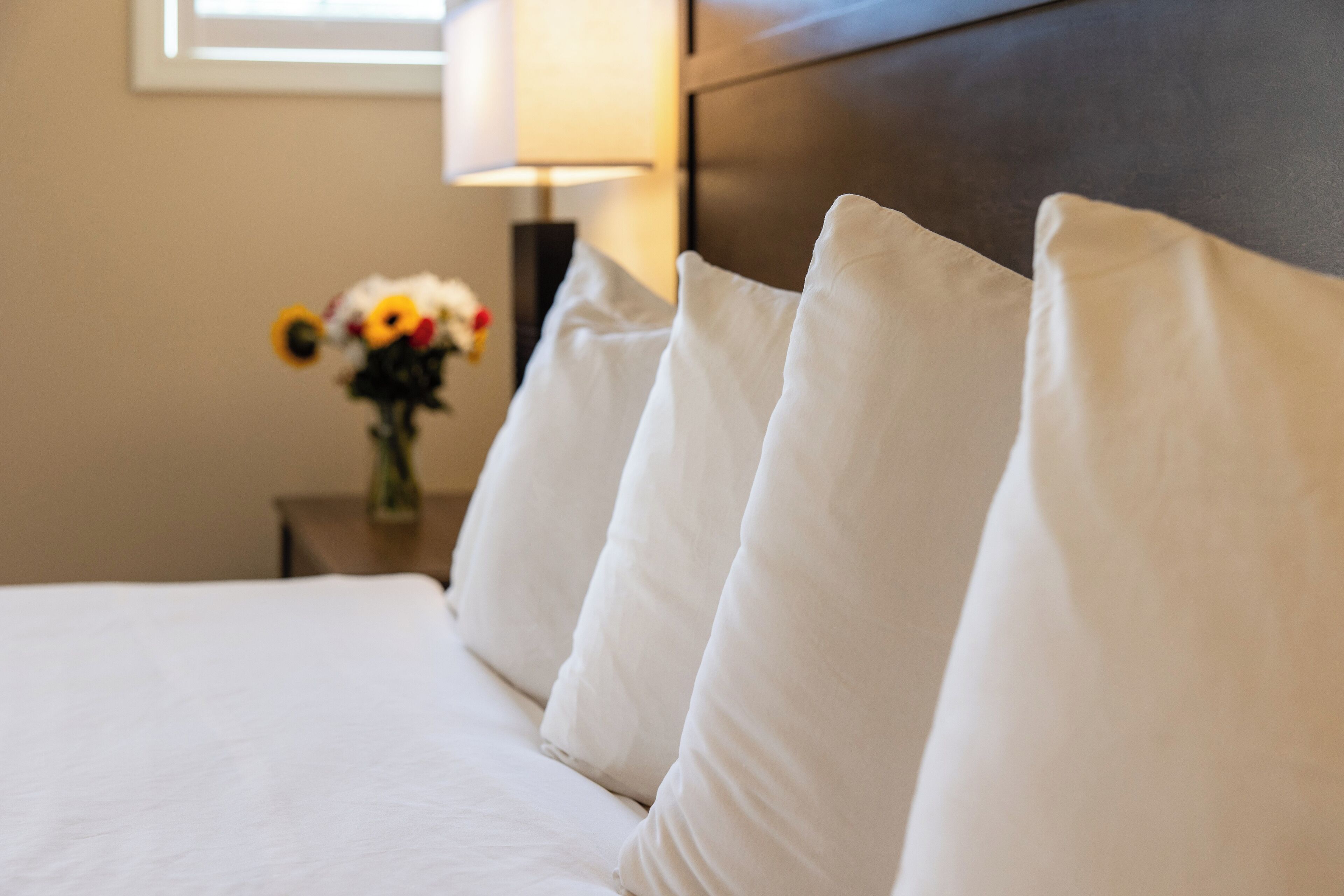 Uma cama bem arrumada com roupa de cama e almofadas brancas, uma mesa de cabeceira com um vaso de flores e uma luminária.
