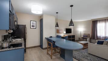 Cocina compacta con una isla central, comedor con mesa y sillas, sofá y televisor en una sala de estar.