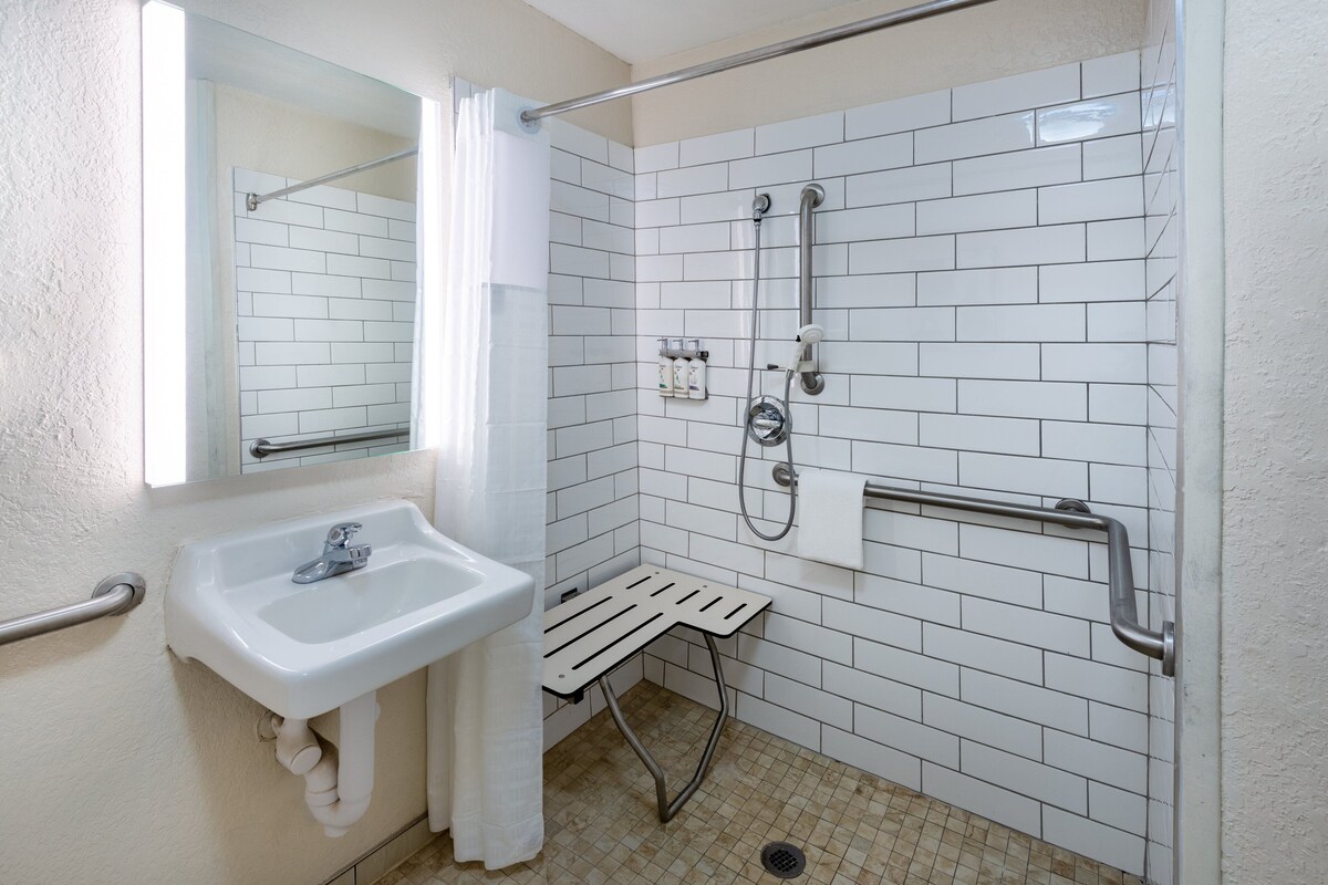 Un baño con lavamanos blanco, un espejo, ducha con barra de apoyo y pared con azulejos.