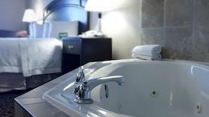 A modern bathroom with a large bathtub, a bedside table with a lamp, and a neatly made bed. - Hampton Inn & Suites by Hilton Edmonton Intl Airport (Leduc)