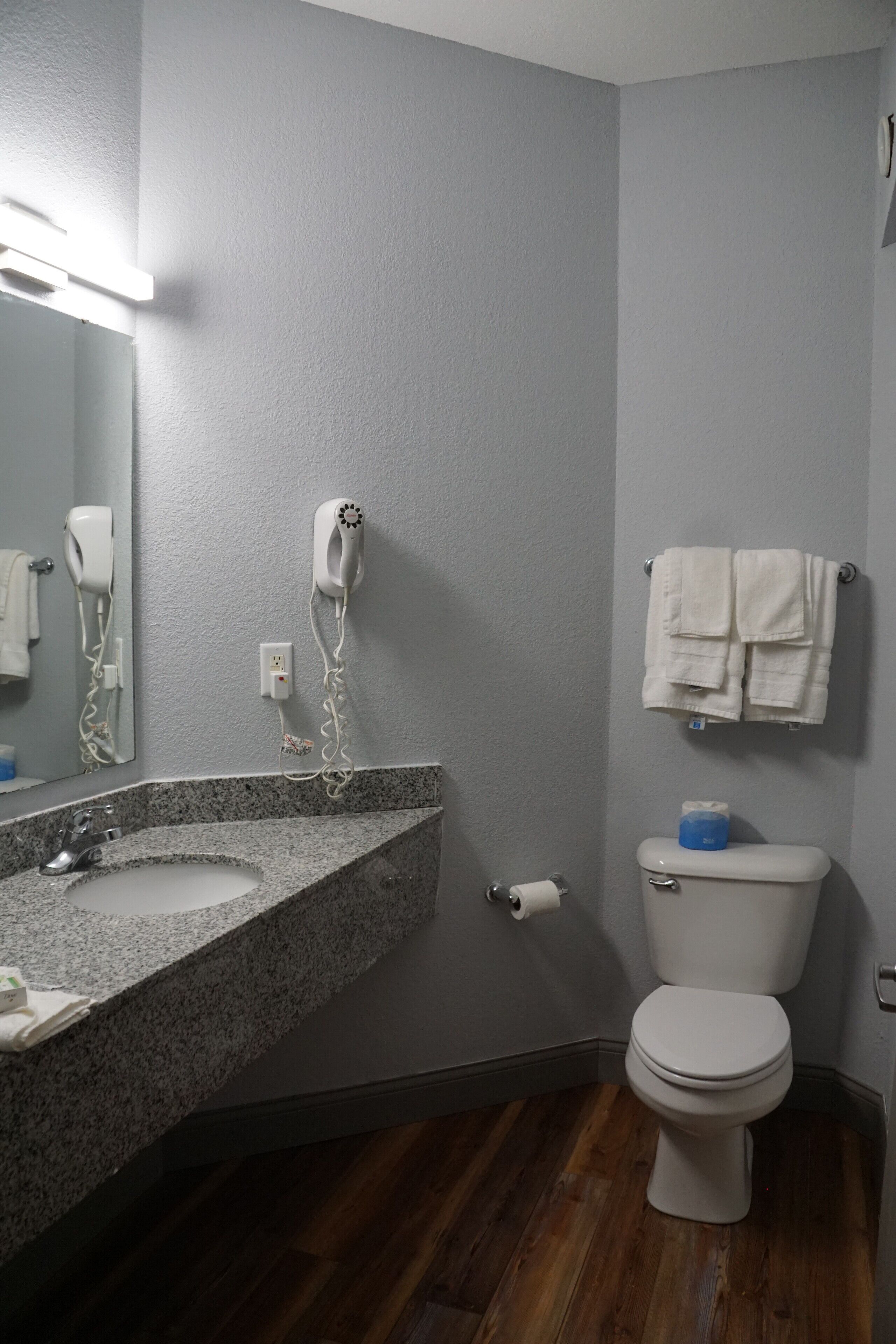 Une salle de bain dotée d’un lavabo avec un plan de toilette en granit, d’un sèche-cheveux fixé au mur, de toilettes avec un porte-papier toilette bleu et d’un porte-serviettes avec des serviettes pliées.