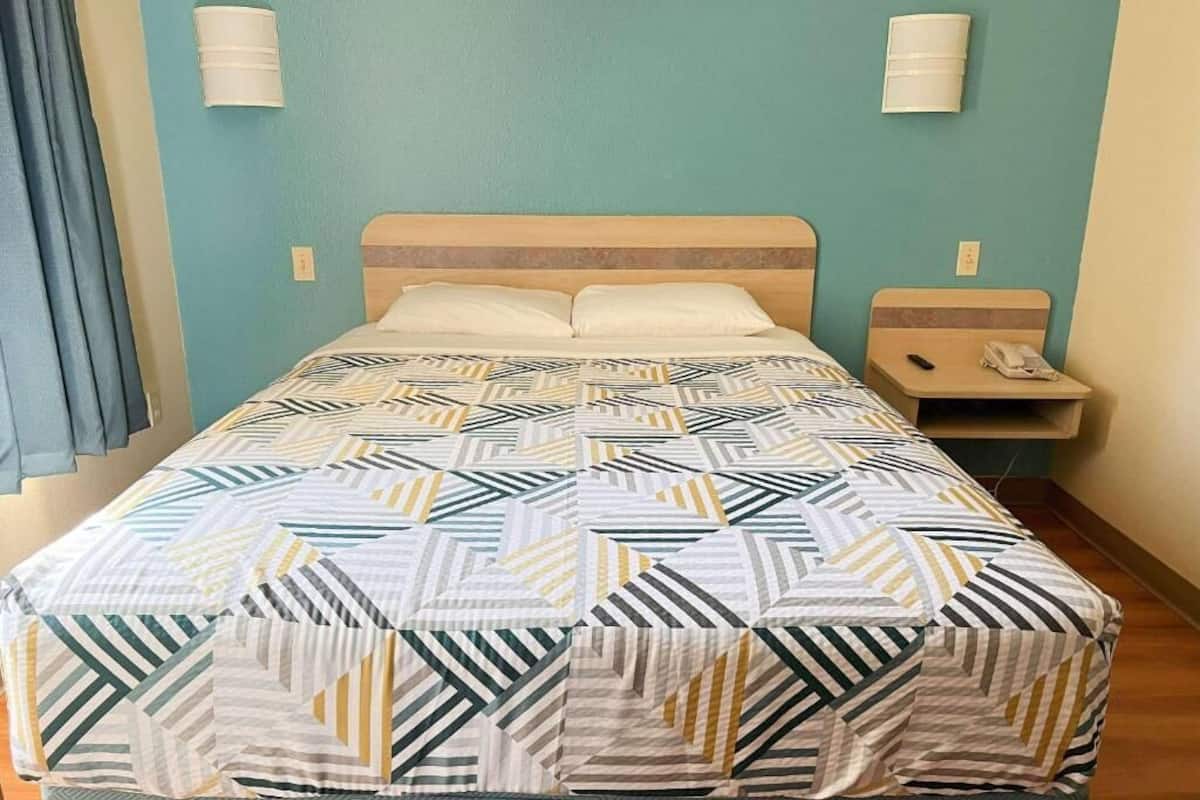 A single bed with a patterned bedspread, a bedside table with a lamp, and a wall-mounted light fixture.