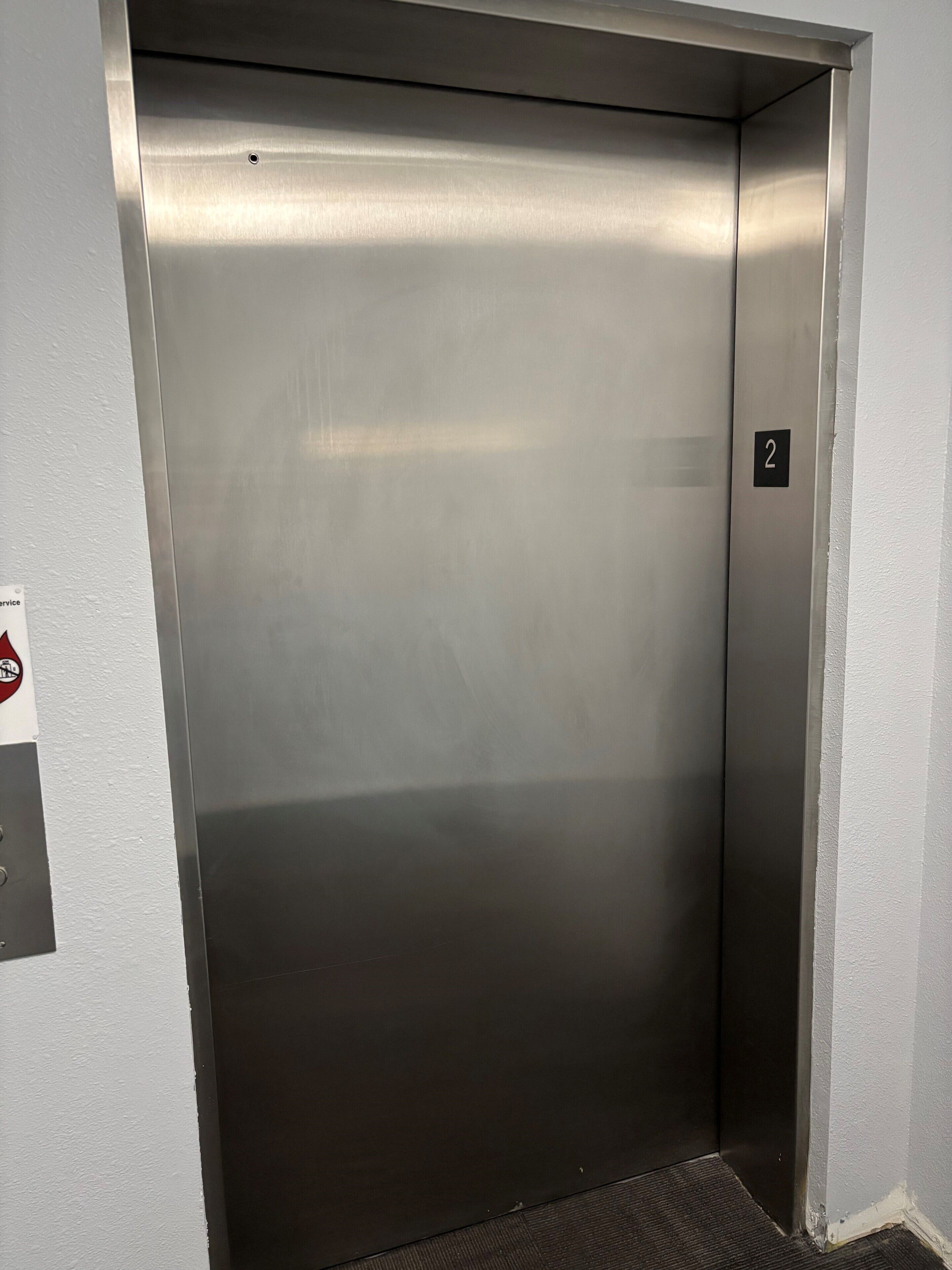 Porta de elevador metálica com placa de identificação com o número 2, tapete de chão texturizado e sinal de serviço na parede.
