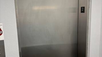 Porta de elevador metálica com placa de identificação com o número 2, tapete de chão texturizado e sinal de serviço na parede.