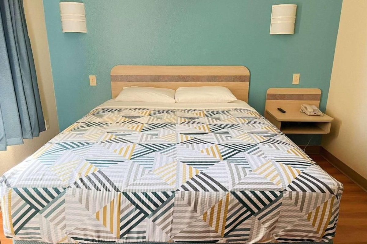 A single bed with a patterned bedspread, a bedside table with a lamp, and a wall-mounted light fixture.