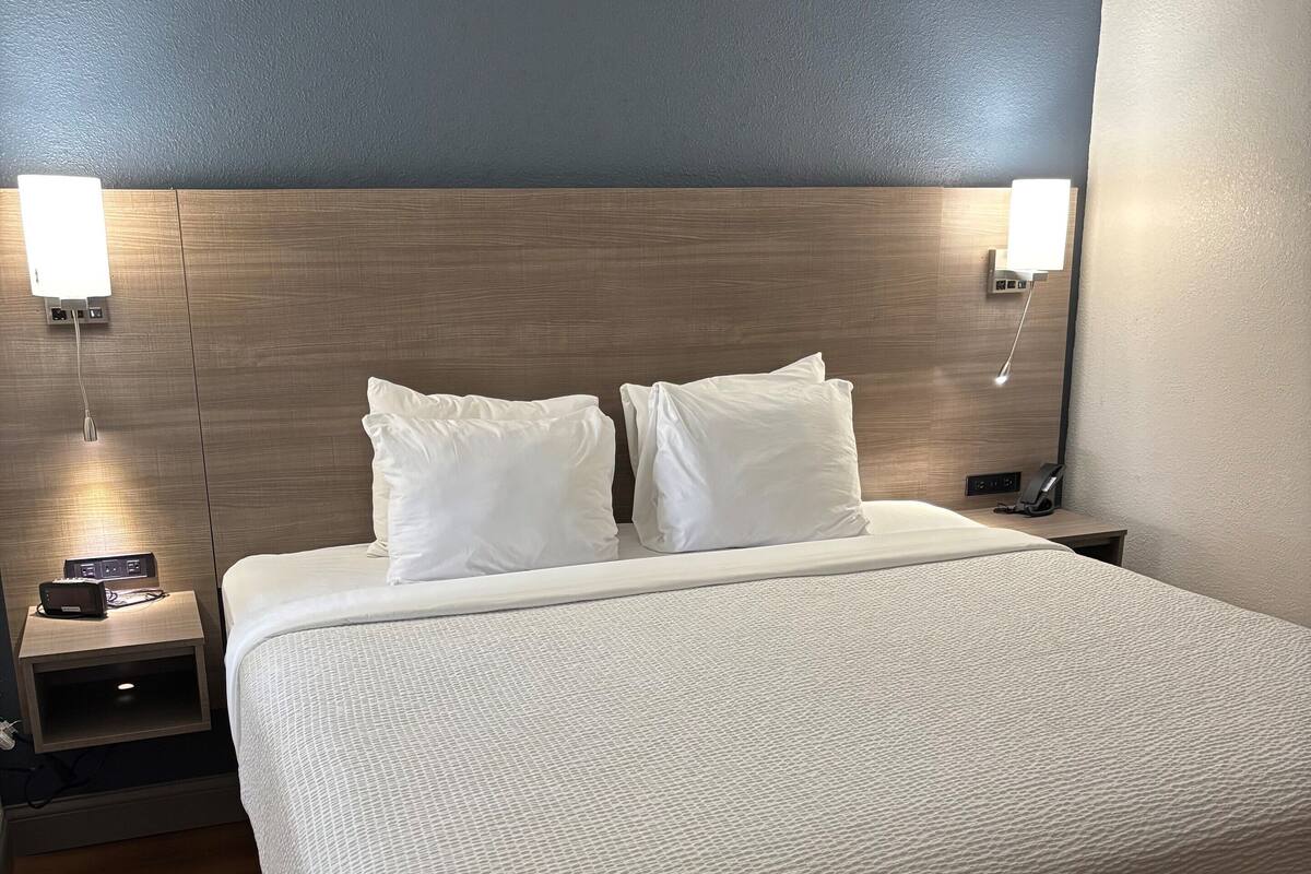 A hotel room with a large bed, two bedside tables, a nightstand, a wall-mounted lamp, and a power outlet.