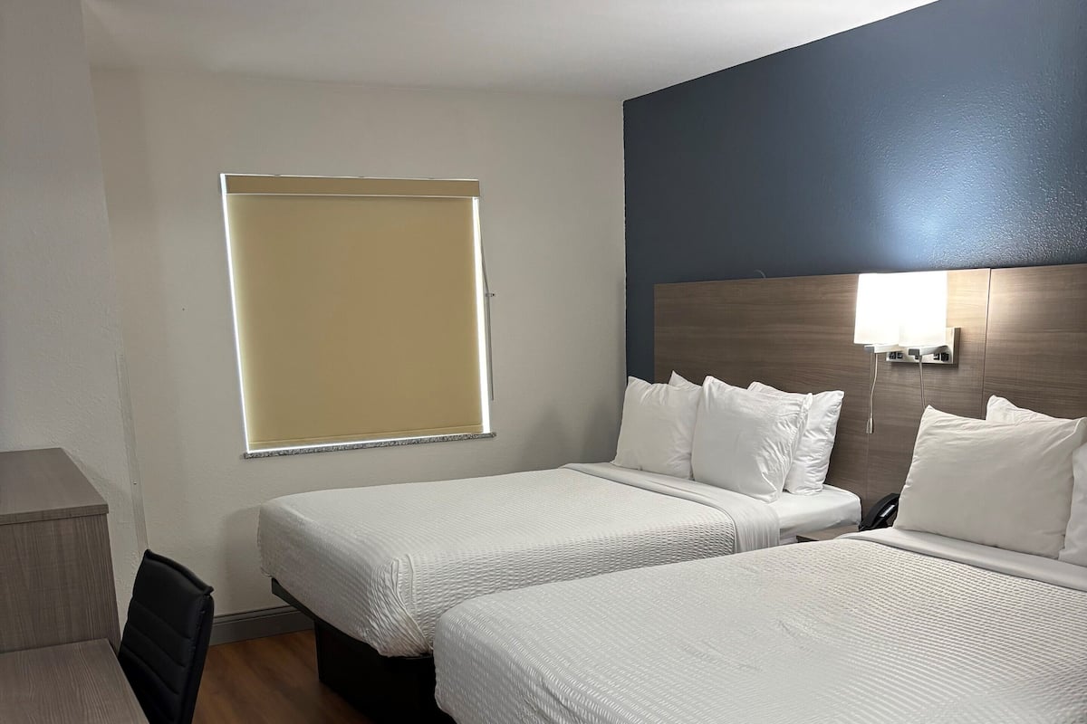 A hotel room with two single beds, a desk, and a window with blinds.