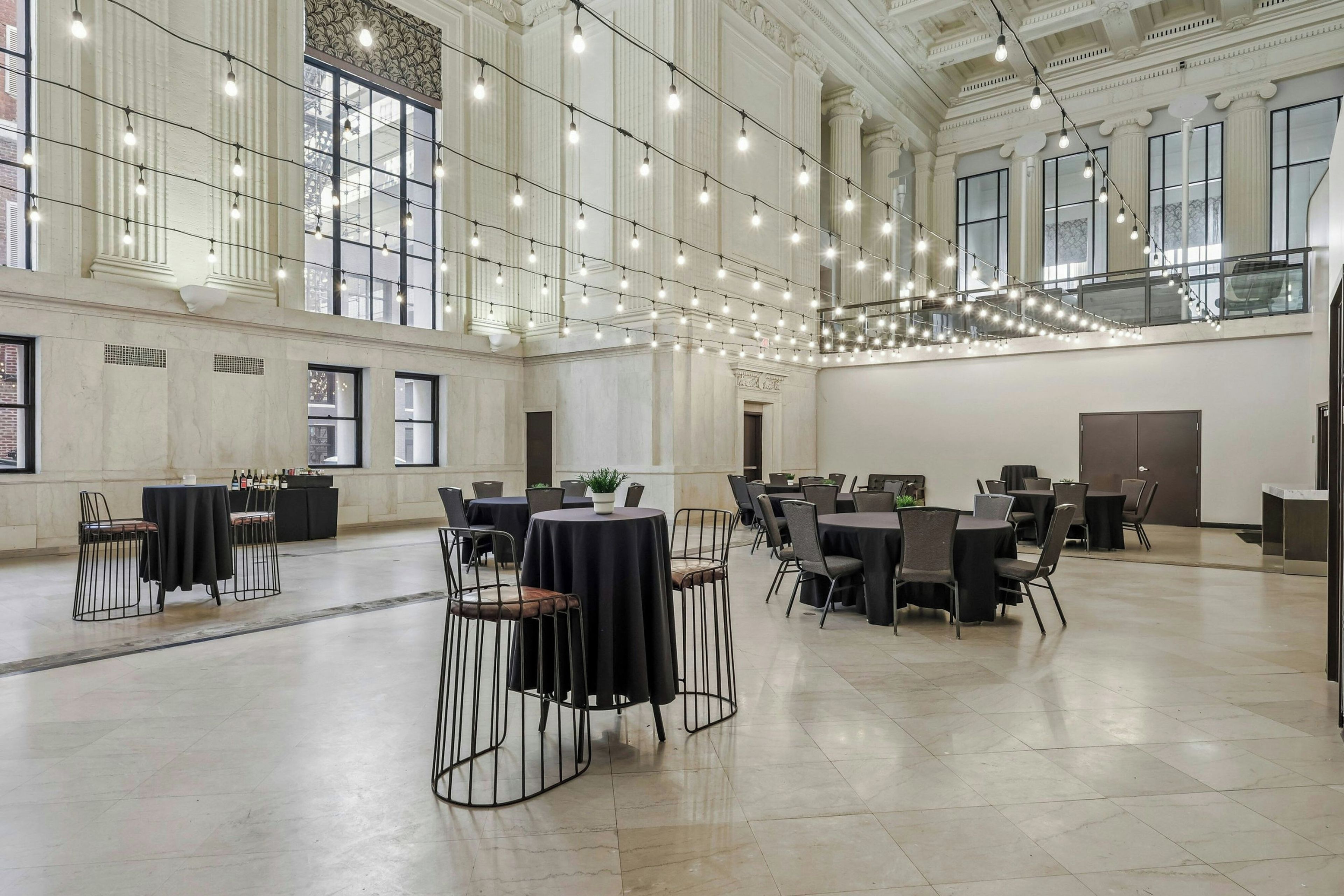 A spacious hall with high ceilings, large windows, and string lights. Tables and chairs are arranged for events.