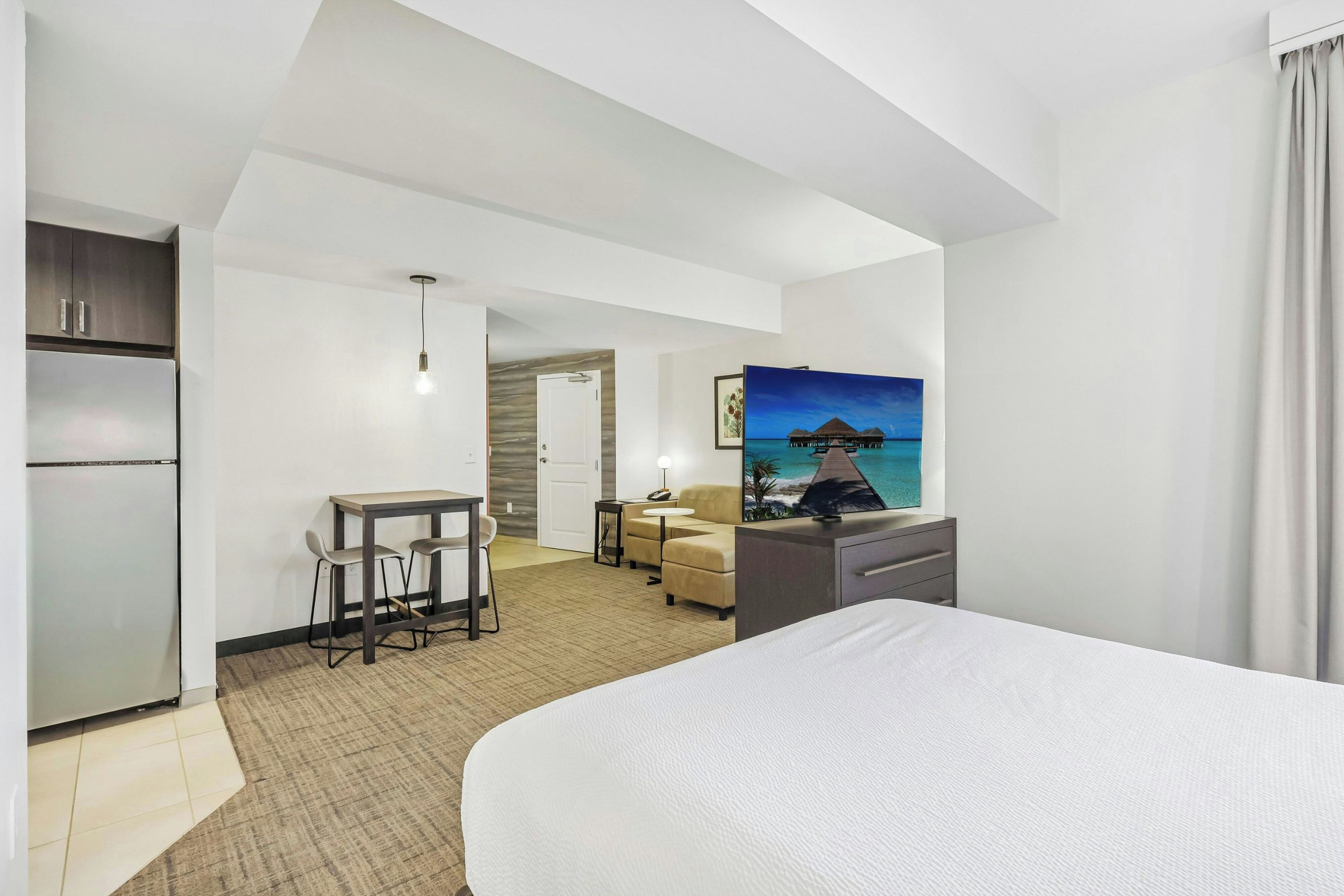 Hotelkamer met een bed, een bureau met stoel, een koelkast, een schilderij en uitzicht op zee.