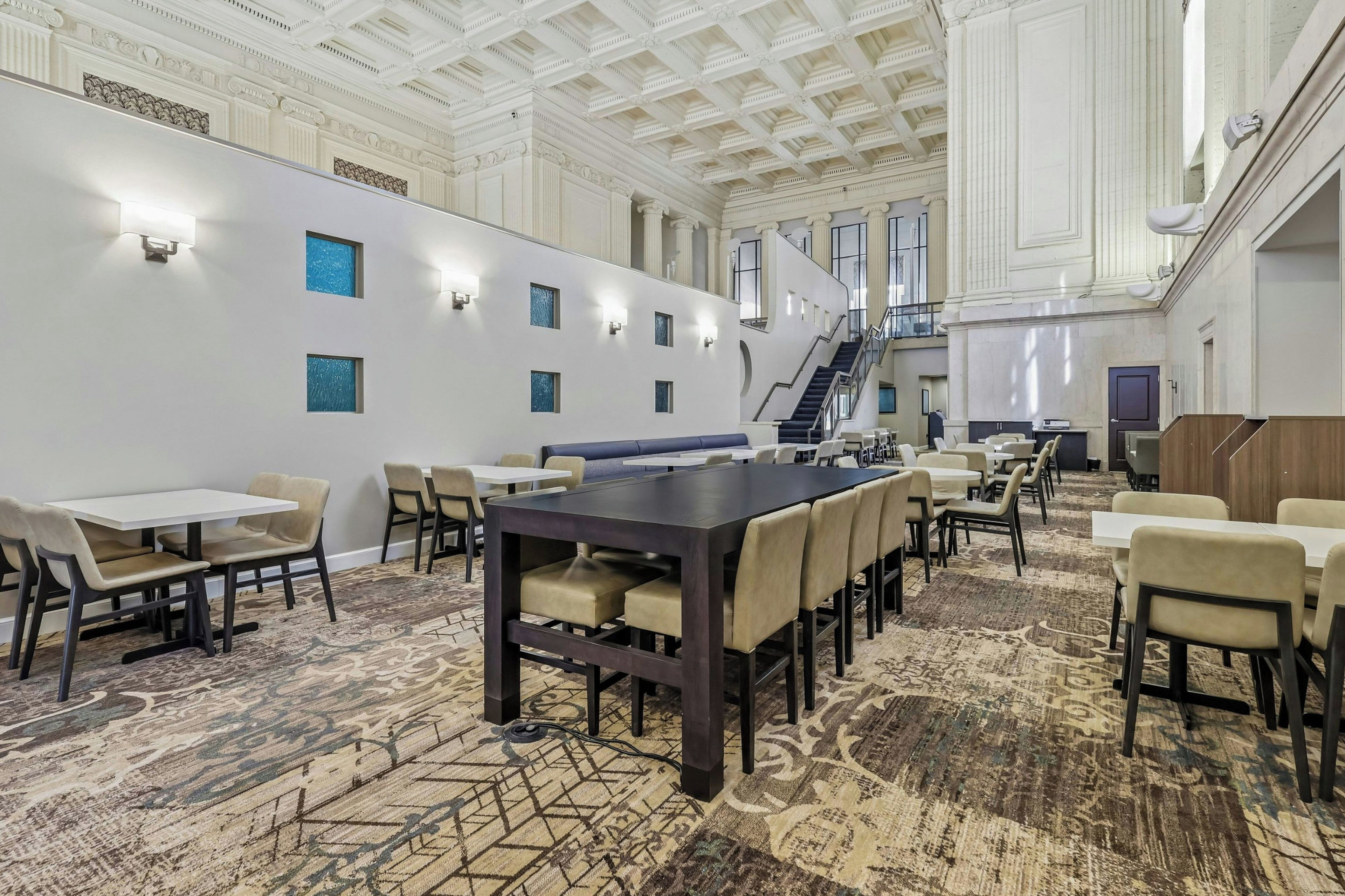 A spacious conference room with a large table and multiple chairs, a staircase, and a high ceiling with decorative moldings.