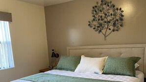 Un lit bien fait, recouvert d’une couvre-lit verte et blanche, avec un élément décoratif accroché au mur au-dessus, et un tapis à motifs sur le sol.