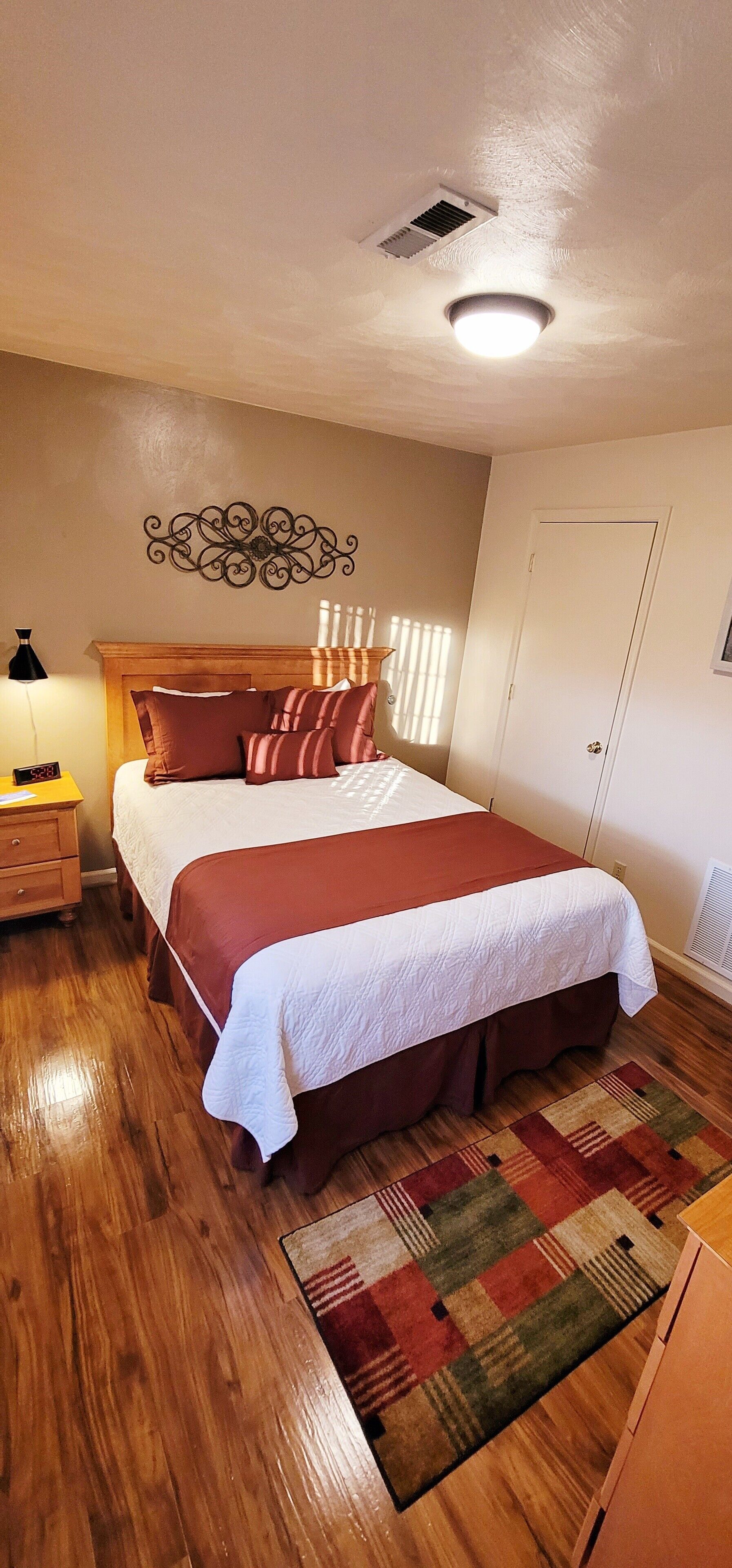 A bedroom with a wooden bed, a nightstand, a wall-mounted air vent, and a patterned rug.