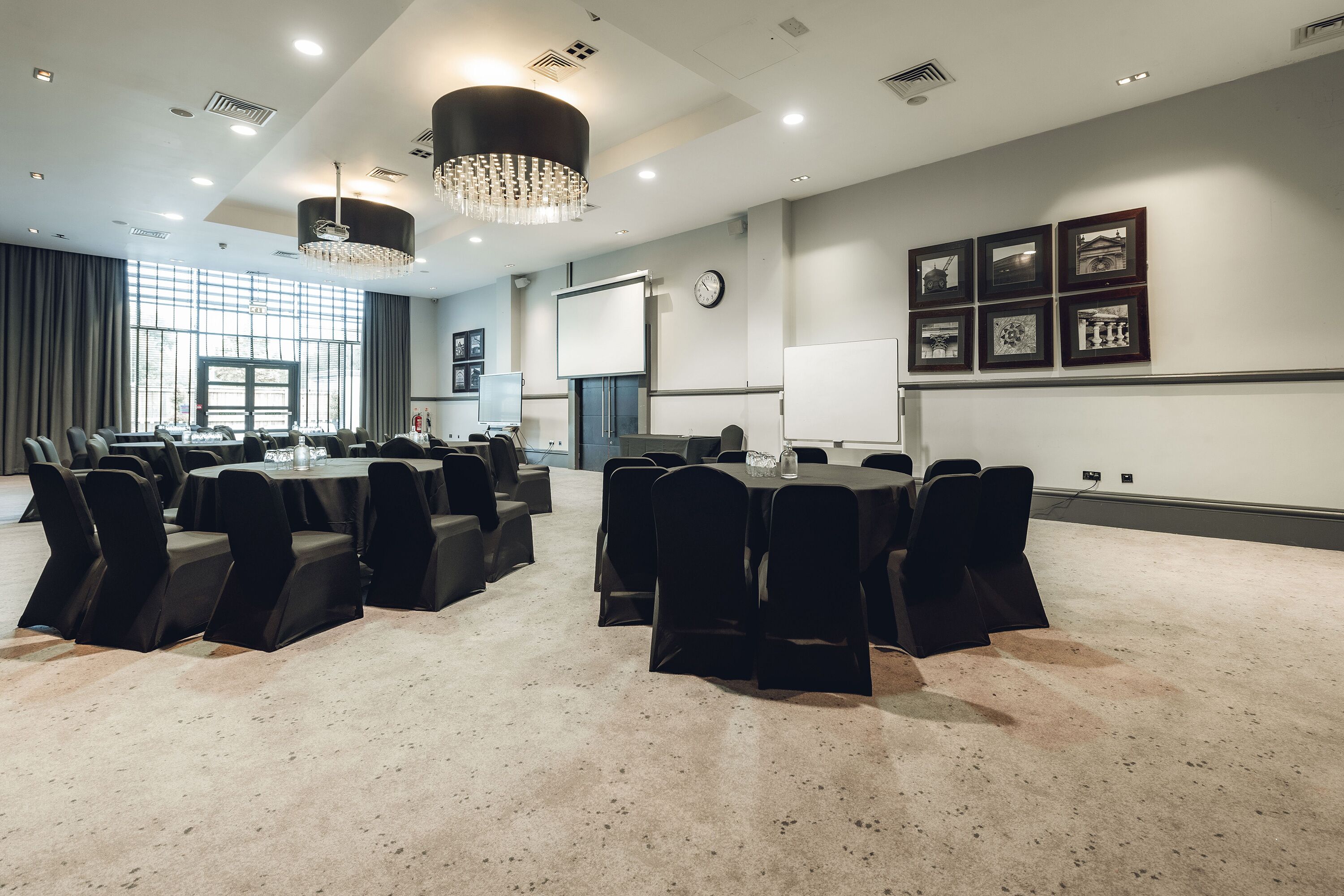 A conference room with multiple tables and chairs, a projector screen, a clock, and framed pictures on the wall.