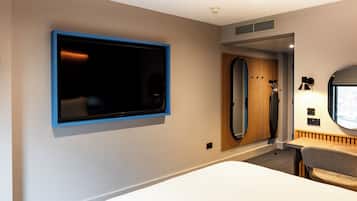 A modern hotel room with a large flat-screen TV mounted on the wall, a wooden panel wall, a round mirror, and a small table with a lamp.