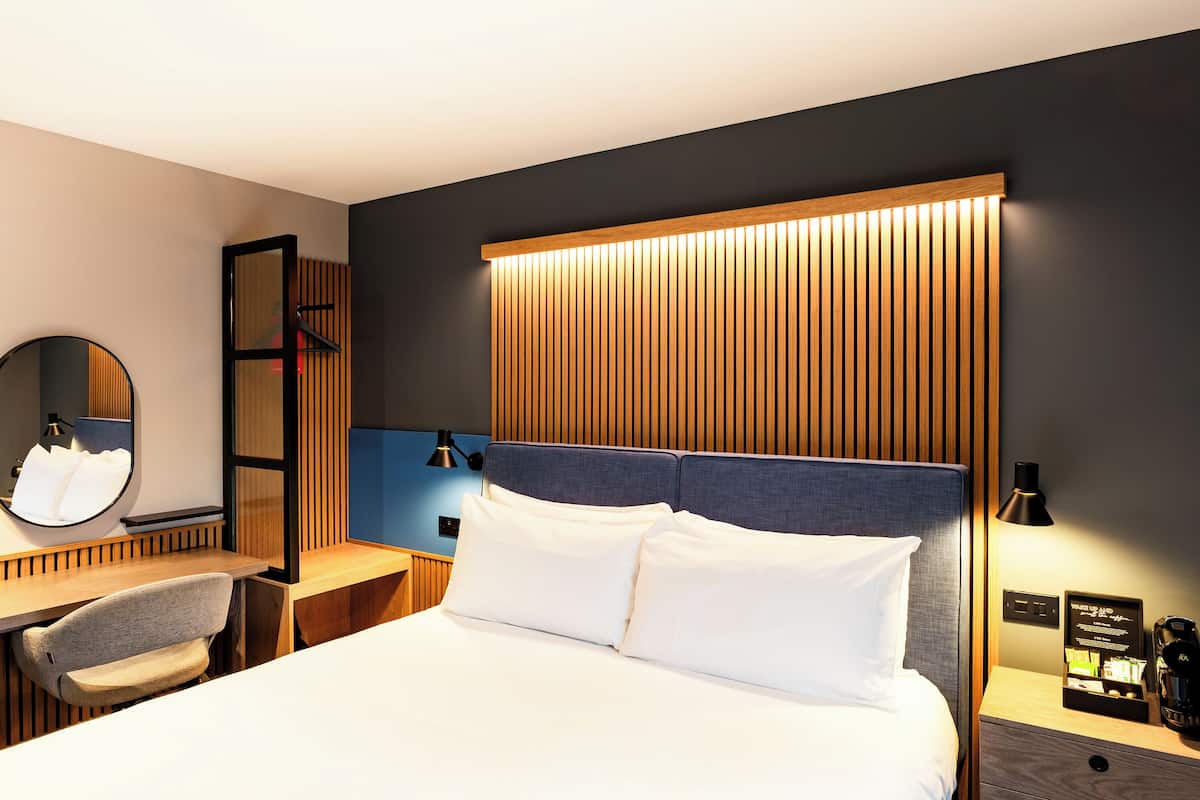 Une chambre d’hôtel moderne avec un grand lit, un bureau avec une chaise, une table de chevet avec une lampe et un miroir.
