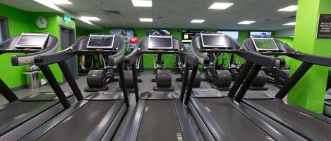 Un gimnasio con varias cintas de correr, paredes verdes y un reloj en la pared.
