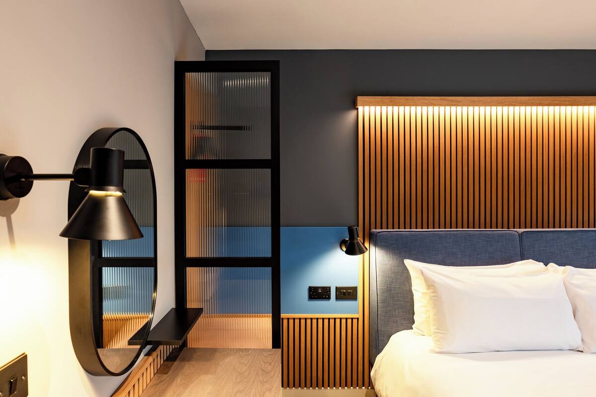 Une chambre d’hôtel moderne équipée d’un lit, d’un bureau, d’une lampe et d’un panneau de commande fixé au mur.