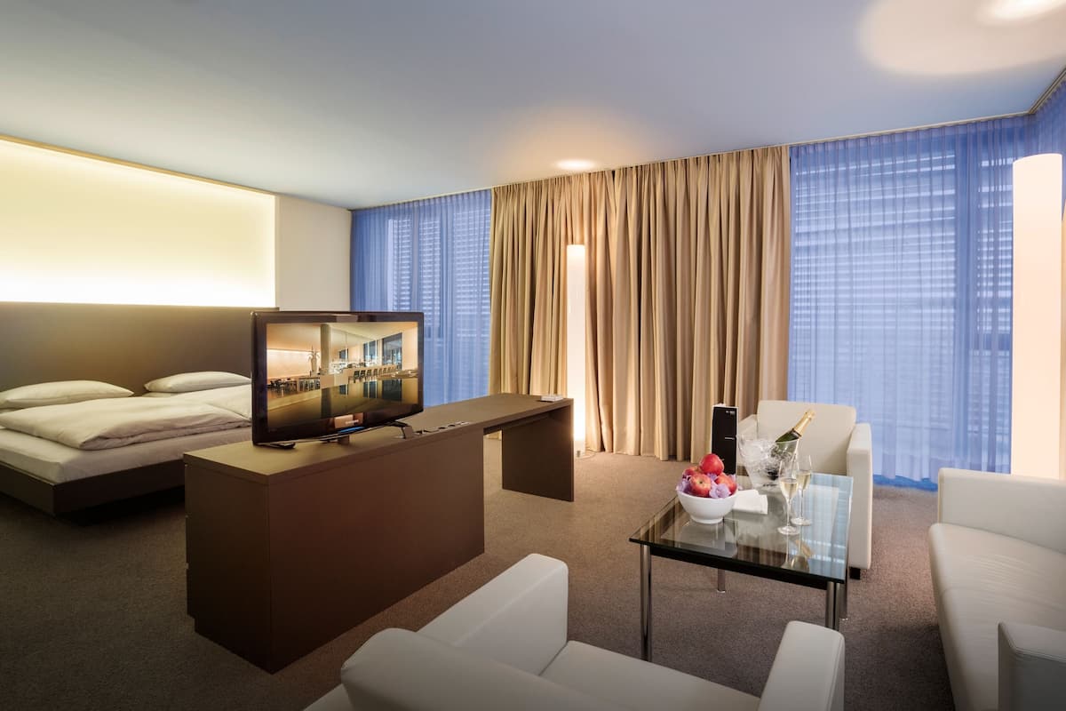 Une chambre d’hôtel comprenant un lit, un bureau avec un téléviseur à écran plat, une table basse en verre avec des fruits et des boissons, un canapé et une vue sur le paysage urbain à travers de grandes fenêtres.