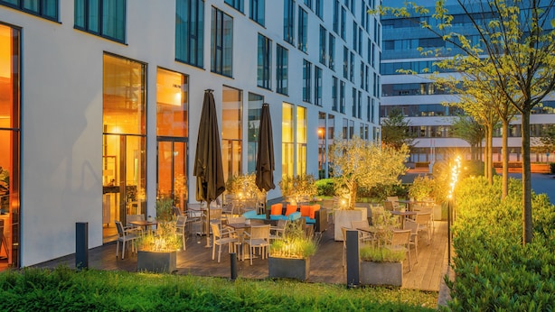A modern outdoor patio area with seating and tables, surrounded by greenery and a building with large windows. - INNSiDE by Meliá Düsseldorf Derendorf (Düsseldorf)